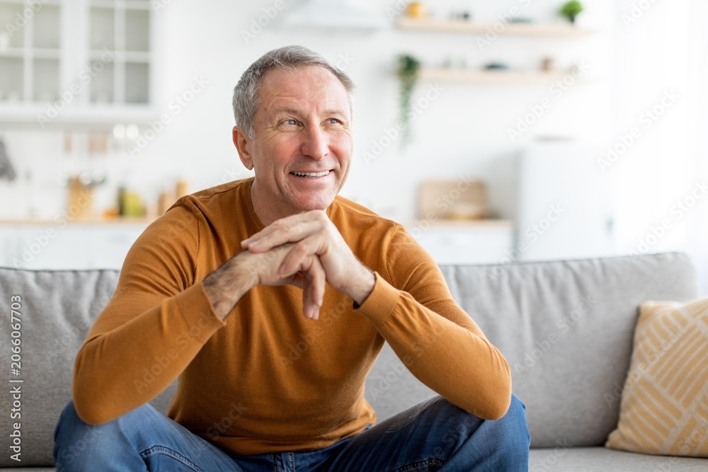 Fototapeta premium Smiling Elderly Caucasian Man Sitting Crosslegged On Couch With Relaxed Emotion At Home In Living Room, Enjoying Leisure Time. Concept Of Aging, Comfort, Relaxation And Lifestyle.