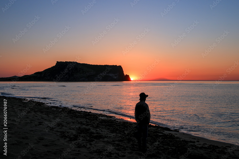 Fototapeta premium A beautiful sunrise over Seongsan Ilchulbong Peak in Jeju Island South Korea. A silhouette of a man stands on the quiet sandy beach looking at the golden sun rising next to the volcanic tuff cone.