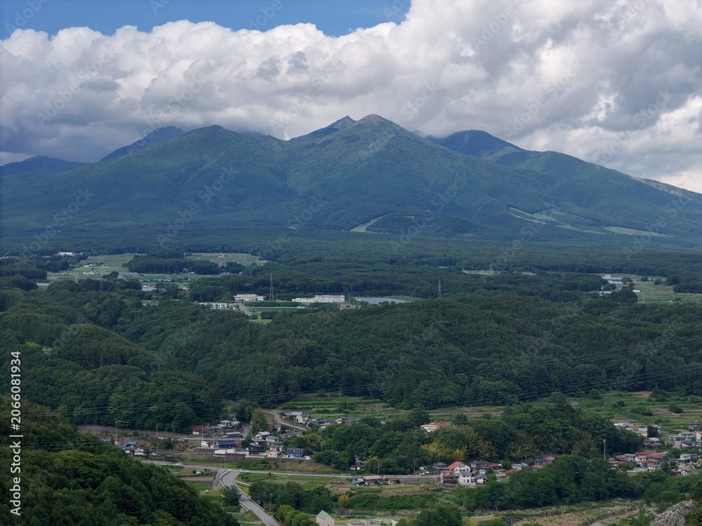 Fototapeta premium Vast mountain range looms above lush green valley. Clouds drift lazily over the peaks. Village sprawls below among trees and fields. Roads wind through quiet countryside