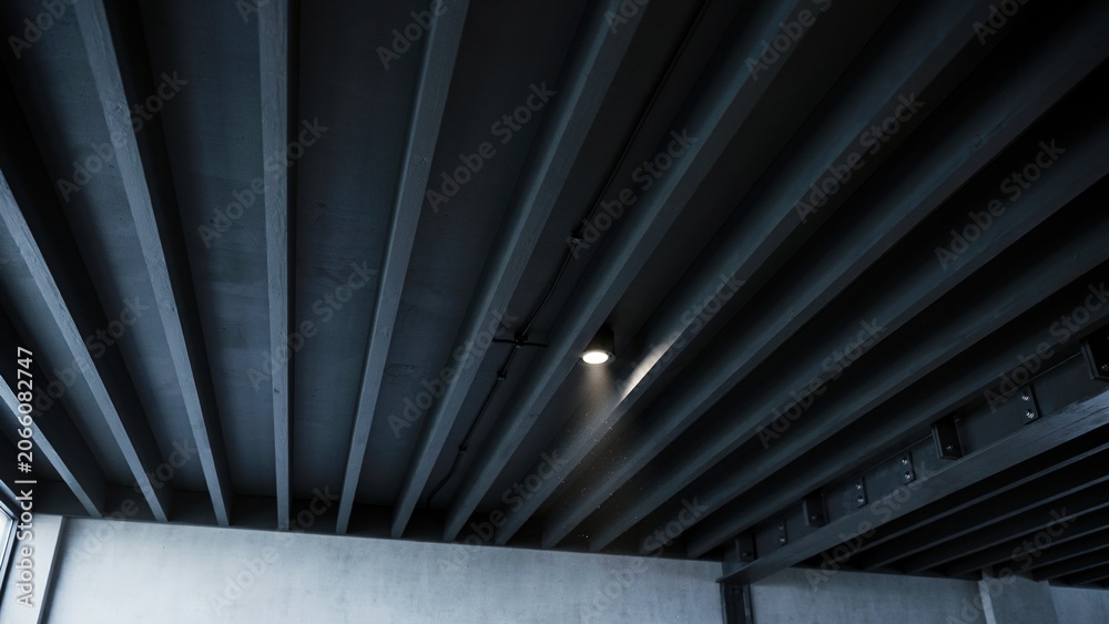 Fototapeta premium Concrete ceiling beams form a dark architectural pattern while a single spotlight casts a bright glow on the grey surface.