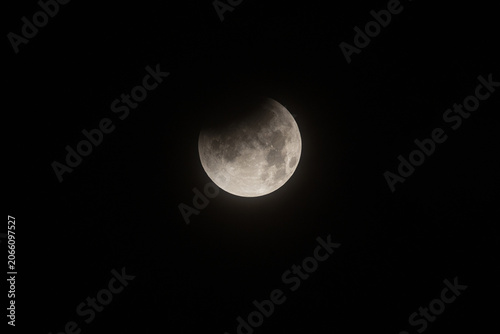 A close up view of a partial lunar eclipse with Earth shadow partially covering the upper dark side of the moon in a black night sky