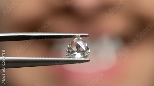 A close-up shot of a diamond held by metal tweezers with a blurred face in the background