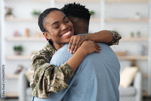 Photography Smiling Black Female Soldier Embracing Her Partner With Joyful Expression At Their Home, Celebrating Reunion After Deployment With Warm Hug, Indoor Setting