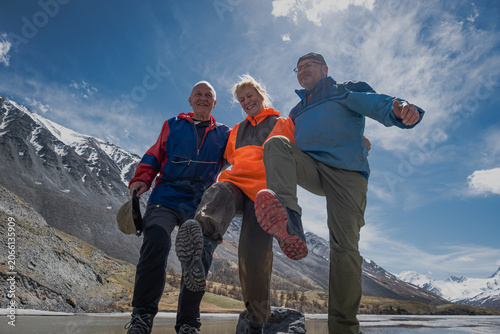 Low angle view of happy senior friends balancing on one leg on frozen lake. Active elderly travelers enjoying extreme trekking travel adventure in scenic mountain wilderness.
