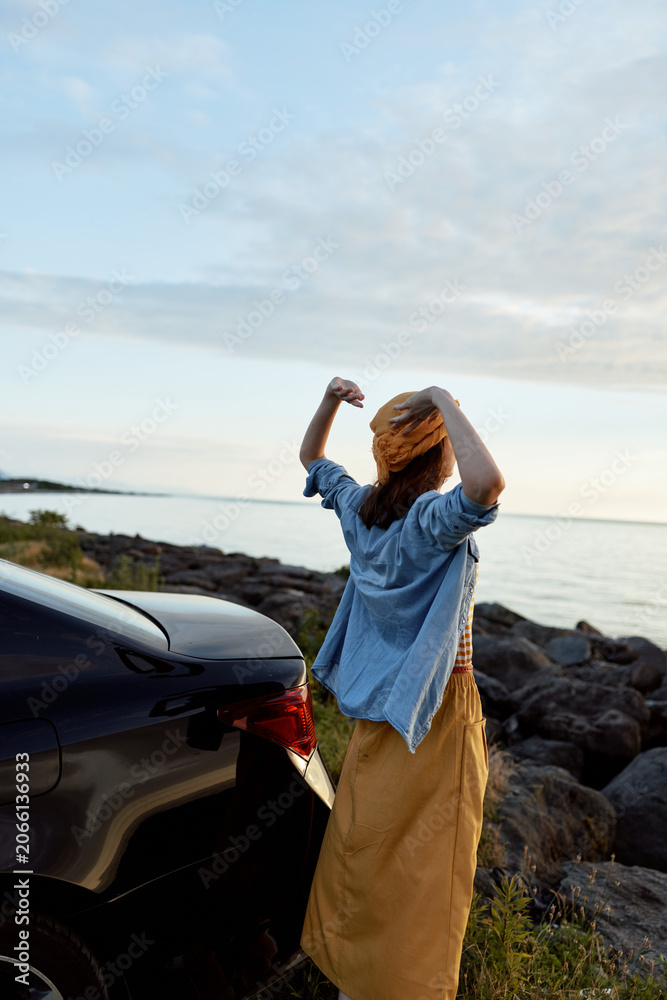 Fototapeta premium A woman stands beside a car on a rocky coast, arms raised in celebration, enjoying fresh air and sunlight. A candid lifestyle moment outdoors.