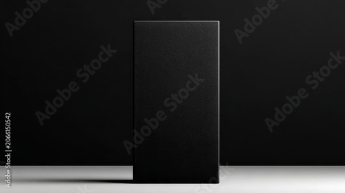a minimalist, tall, black rectangular box centered against a dark background, resting on a light-colored surface with a soft shadow.