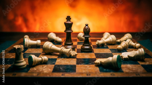 Tableau sur toile Dark wooden king and bishop stand defiantly at the center of a rustic chessboard, surrounded by defeated white pieces against a fiery orange background