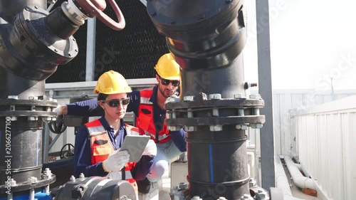 Two industrial technicians wearing helmets and safety gear inspecting and adjusting a large valve on mechanical pipeline infrastructure.