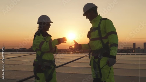 Two solar technicians in safety gear shaking hands on rooftop photovoltaic panels during sunset, symbolizing successful renewable energy project completion.