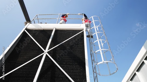Two industrial technicians climbing and working on a ladder and platform of a large cooling tower structure at an industrial facility.