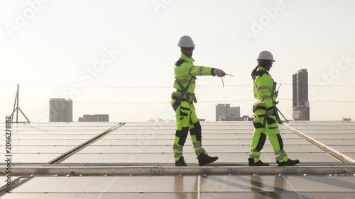 Two solar technicians sitting on rooftop photovoltaic panels at sunset discussing inspection results and pointing toward the solar installation area.