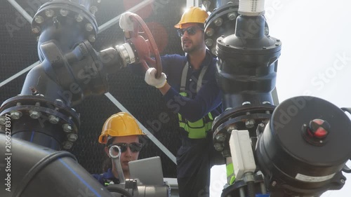 Two industrial technicians wearing helmets and safety gear inspecting and adjusting a large valve on mechanical pipeline infrastructure.