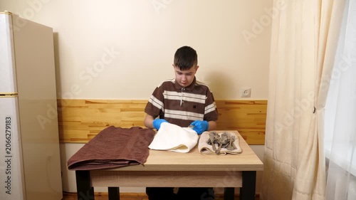 Teenager wearing gloves polishes cutlery with a towel at a table, carefully preparing clean utensils for use.