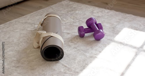 Rolled mat and dumbbells are resting on rug with camera shift moving sunlight, signaling workout