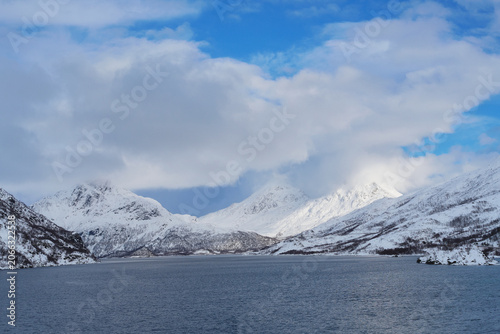 Scenic Winter Lofoten Road Trip View of Snow-Capped Mountains Meeting Deep Blue Fjord Water Under Cloudy Sky in Arctic Norway, Majestic Nordic Landscape Photography.