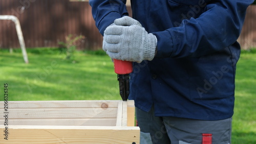 Fotografía Close-up of worker in blue uniform and safety gloves using a red pneumatic nail gun to assemble a wooden structure outdoor