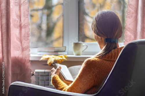 Foto Woman is reading book while sitting in an armchair by window with cup of coffee
