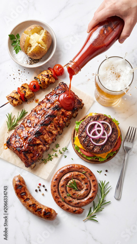 Flat lay of grilled BBQ food on a white marble background. A hand pours red ketchup onto delicious ribs next to a hamburger, sausages, skewers, baked potato, and splashing beer glass.