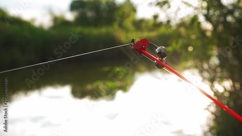 Fishing rod tip with bell bite indicator shaking during fish bite near riverbank at sunset. Concept of recreational fishing and successful catch
