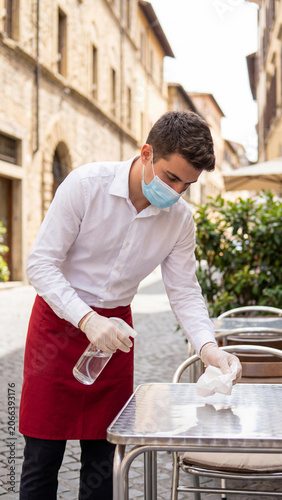 A waiter in a protective face mask and gloves cleaning an outdoor cafe table with disinfectant spray. Post-pandemic safety protocols, restaurant hygiene, hospitality industry, new normal.