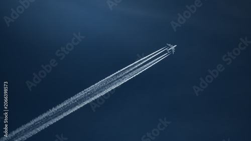 Airplane high in dark blue sky with long white streaks of exhaust fumes behind it