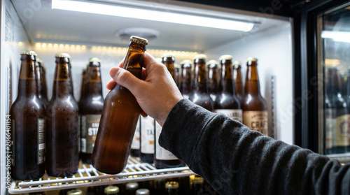 Person hand picking cold beer bottle from refrigerator shelf in store. Close up of grabbing fresh beverage for refreshing drink.