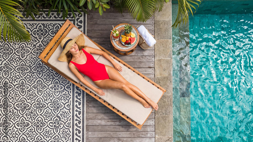 Overhead top view of a young Caucasian woman in a red swimsuit and straw hat relaxing on a wooden lounger by a turquoise pool. Luxury tropical resort villa backyard on a sunny summer day.