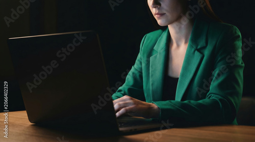 Businesswoman working on laptop at night, focused professional typing on computer in dark office with green blazer.