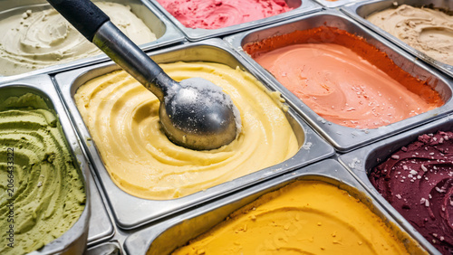 Metal scoop resting over colorful ice cream trays in frozen dessert display with variety of creamy gelato flavors arranged in counter containers at shop, sweet refreshment and dessert selection