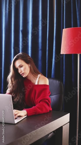Focused Young Woman with Long Brown Hair, Wearing an Off-the-Shoulder Red Sweater and Earbuds, Concentrated on Her Laptop at a Dark Wood Desk with a Red Floor Lamp Against Blue Curtains, Home Office