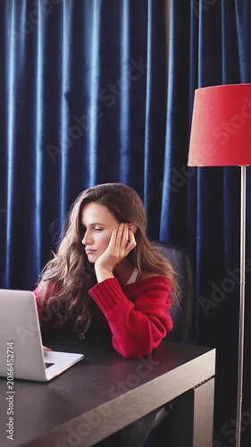 Exhausted and Sad Woman on Laptop at Home Office, Leaning on Her Hand Feeling Fatigued, Sadness and Loneliness Concept, Tired Brunette Freelancer Working Late at Desk Near Red Lamp and Blue Curtains