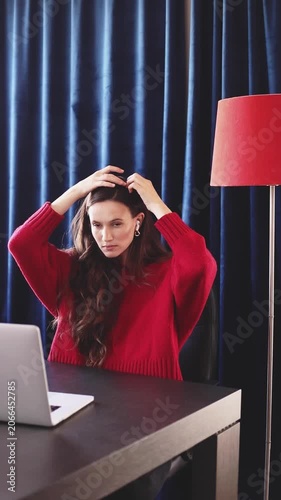 Attractive Young Woman Adjusting Hair and Red Sweater Before Video Call on Laptop, Preening and Flirting at Home Office Desk with Red Floor Lamp Against Dark Blue Curtains, Freelancer Preparation