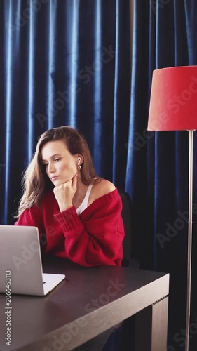 Focused Young Woman with Earbuds Translating Text on Her Laptop, Turning Head and Checking Documents closely at Dark Wood Desk with a Red Floor Lamp and Blue Curtains Background, Translation Work