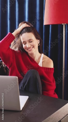 Smiling young woman in red sweater enjoying pleasant conversation on a laptop, professional remote work concept, cozy home office, freelancer lifestyle, authentic video.