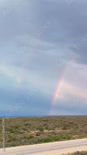 Vertical drone footage sliding with the road across the Nullarbor bushland under a vivid double rainbow