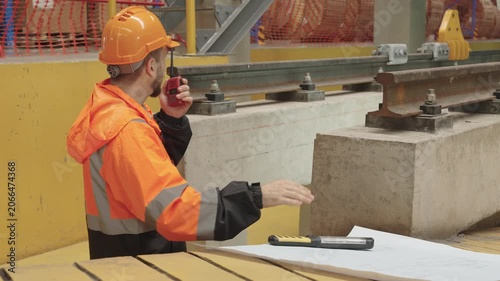 Serious construction worker intently studies detailed bridge blueprint amidst active building site operations, exhibiting focused engineering work and meticulous planning near wooden surface.