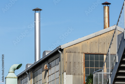 Rustic Japanese industrial factory building with rain-capped exhaust chimneys, corrugated metal siding, and multi-pane industrial windows set to a clear blue sky.