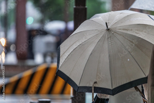 Obraz na plátne Large black‑trimmed beige canopy shelters a passerby at a busy intersection, with an orange‑black striped barrier prominetly visible in a bokeh background of soft lights and indistinct figures