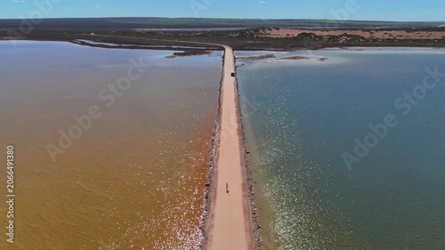 Aerial drone shot rising while flying forward along the straight road through Point Sinclair Pink Lake with bright sun reflections on the water