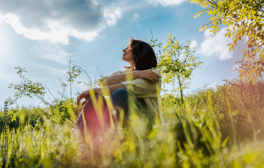 Fototapeta premium Low angle shot of a dreamy middle aged woman sitting on green grass against a beautiful sky with clouds