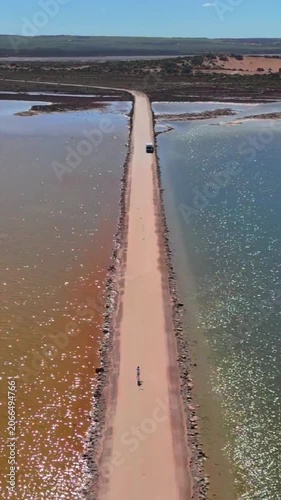 Vertical aerial drone shot rising while flying forward along the straight road through Point Sinclair Pink Lake with bright sun reflections on the water