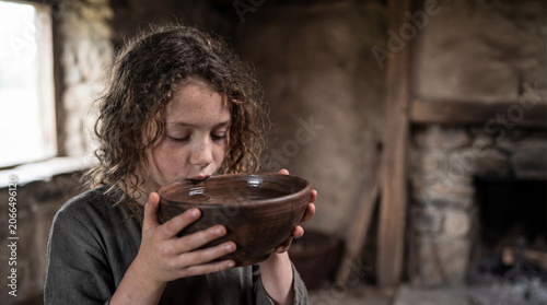 A young girl with curly hair holding and drinking from a rustic clay bowl inside a dim historic stone room at daytime