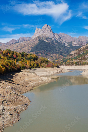 An autumnal landscape of a valley with a beautiful mountain and, at its foot, a reservoir that has been drained to collect snow and water for the winter.