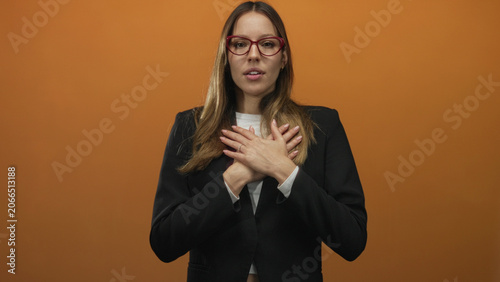 Fotografija Woman with hands crossed over chest in studio orange set, wearing red glasses and a black blazer, looking sincere; gratitude sincerity calm
