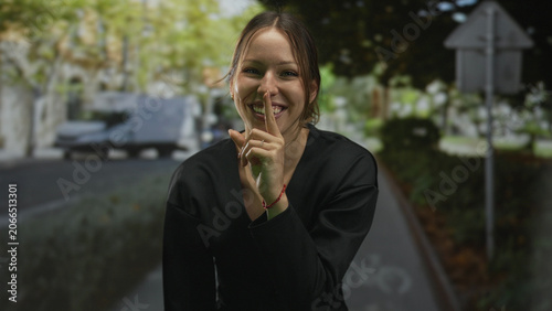 Fotografie Young hispanic woman smiling, finger to lips for silence on a city street with bike lane and trees; playful secrecy