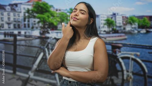 Canvas Print Young woman touching temple beside bicycle on canal street, wearing white tank top and jeans while smiling slightly; confidence thinking