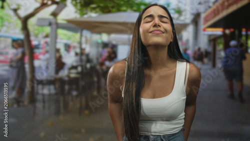Fotografija Hispanic woman in white tank top with eyes closed smiling and leaning forward at street cafe, showing bare shoulder and long hair; contentment