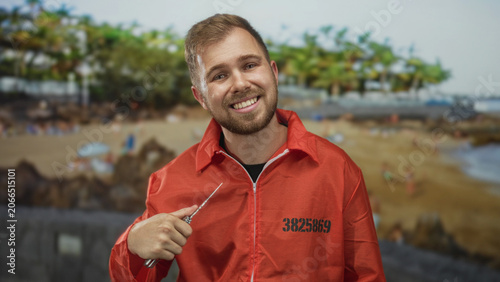 Foto Man in orange prisoner jumpsuit with visible inmate number smiling while holding a pocketknife near his chest on a street by the beach; mischief