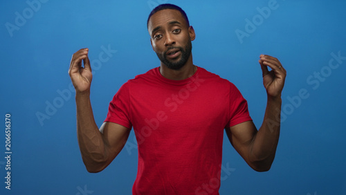Young man in a red shirt raising both hands with palms visible against a blue studio wall; thoughtful reflection.
