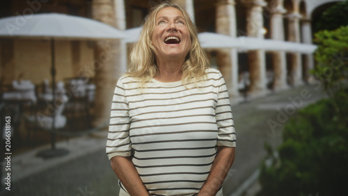 Obraz na plátně Senior woman with hands clasped looking down in front of building courtyard, striped shirt visible and subtle smile; quiet contemplation